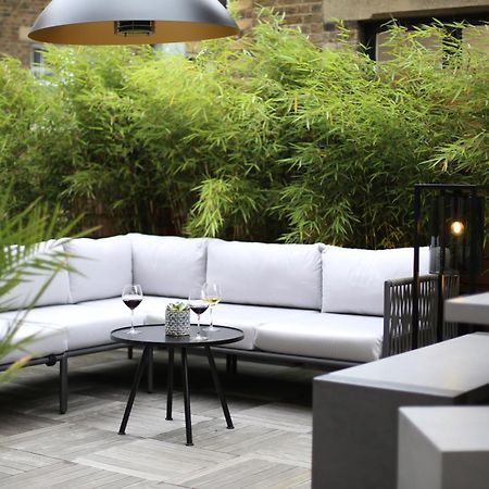 Middle Eight - Covent Garden - Preferred Hotels And Resorts 伦敦 外观 照片 The photo shows an outdoor seating area featuring a modern gray sectional sofa with plush cushions. In front of the sofa is a small, round black table with two glasses of wine on it, accompanied by a decorative plant or vase. Surrounding the area are