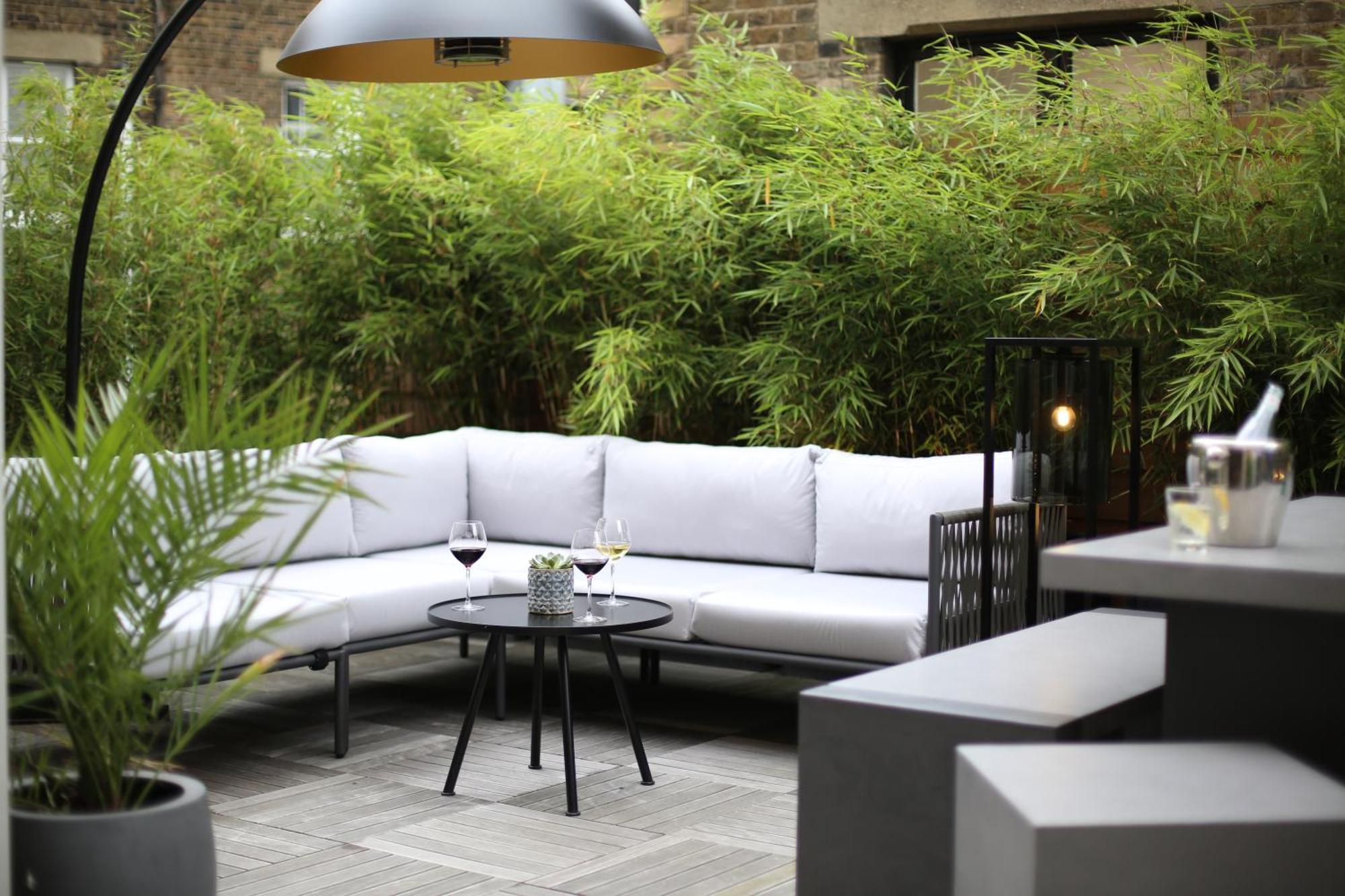 Middle Eight - Covent Garden - Preferred Hotels And Resorts 伦敦 外观 照片 The photo shows an outdoor seating area featuring a modern gray sectional sofa with plush cushions. In front of the sofa is a small, round black table with two glasses of wine on it, accompanied by a decorative plant or vase. Surrounding the area are